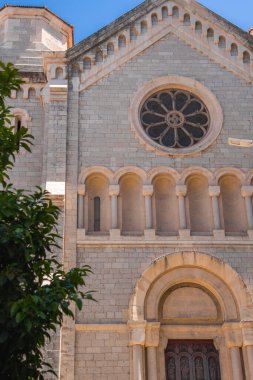 Cannes 'daki Romen katedralinde bir gül penceresi, kemerli pencereler ve bir ağaç ve parlak mavi gökyüzü tarafından çerçevelenmiş görkemli bir giriş yer alıyor..
