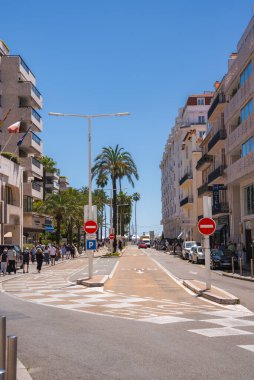 Cannes, Fransa 'da canlı bir cadde, içinde palmiye ağaçları, modern ve klasik binalar, görünür bir otel, yayalar, park edilmiş arabalar ve uzak bir yat var..