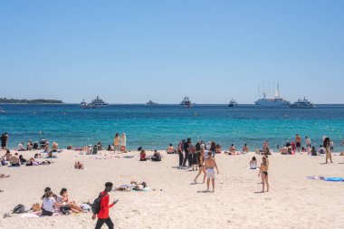 Fransa, Cannes 'da kumsalda dinlenen insanların olduğu hareketli bir plaj. Turkuaz Akdeniz 'de yatlar, bir yelkenli ve bir ağaç çizgili ufuk çizgisi bulunur..