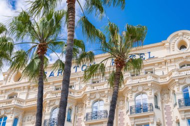 Fransa 'nın Cannes kentindeki Carlton Oteli' nde parlak mavi gökyüzünün altında süslü Belle Epoque tasarımı, krema cephesi, kemerli pencereler ve palmiye ağaçları yer almaktadır..