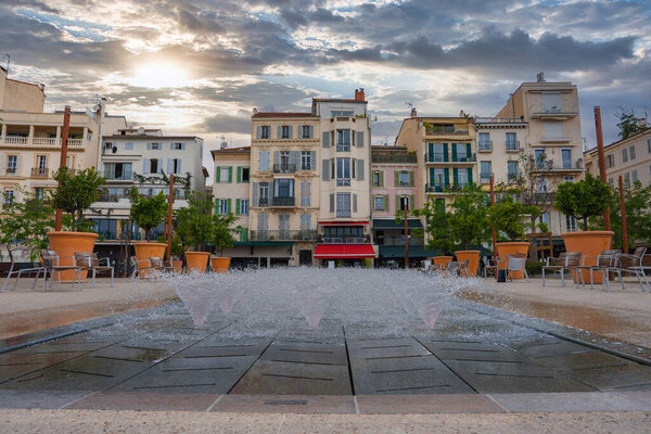 A square in Cannes, France, features a fountain with water jets, pastel colored buildings with shutters, potted trees, and outdoor seating under an overcast sky.