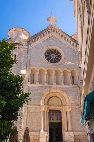 Fransa 'nın Cannes kentindeki Romen katedralinde bir gül penceresi, kemerli kapılar ve yakınlardaki binalar ve bir ağaç tarafından çerçevelenmiş ayrıntılı taş işçiliği yer alıyor..
