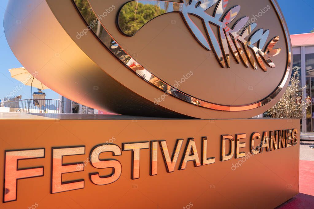 Golden Festival de Cannes sign with logo, reflecting trees and outdoor seating. Red carpet in the background suggests a sunny day in Cannes, France.