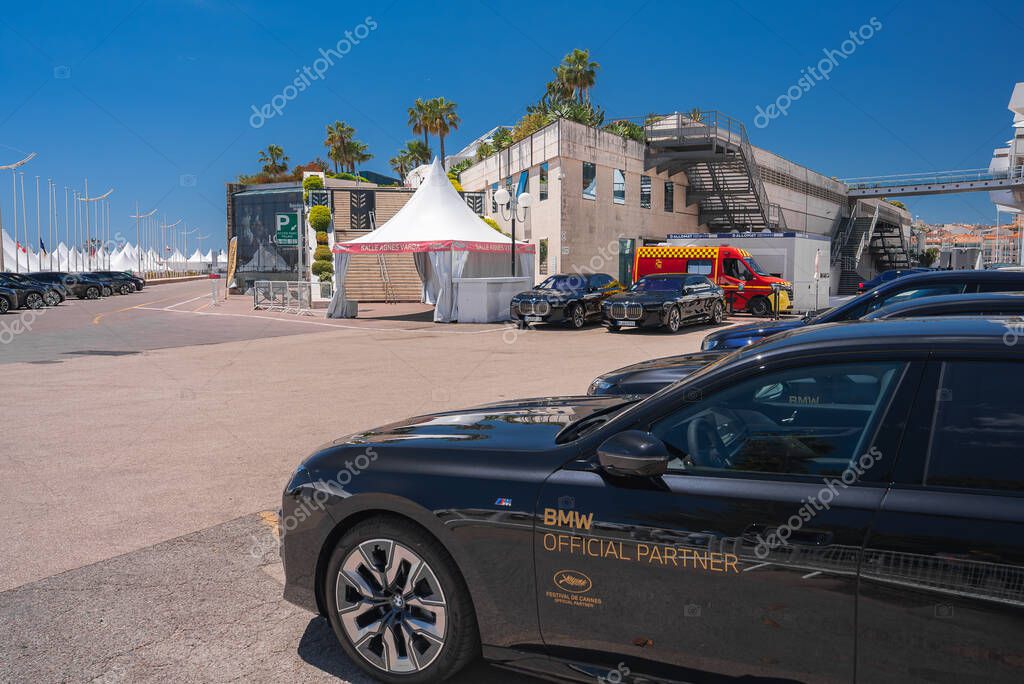 A black BMW with Cannes Film Festival branding is parked near a modern building with stairs, white tents, luxury cars, palm trees, and a clear sky.
