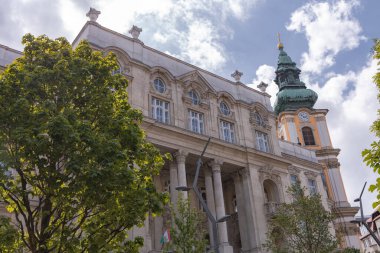 Budapeşte 'deki büyük tarihi binada kemerli pencereler, süslü taş işçiliği ve ağaçlar ve bayrak direğiyle çerçevelenmiş altın kubbeli yeşil kule yer alıyor..