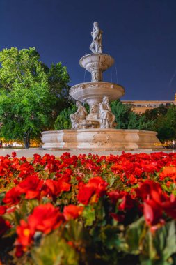 Budapeşte 'de çok katmanlı taştan bir çeşme. Etrafı kırmızı çiçekler, yeşil ağaçlar ve açık bir gökyüzünün altında aydınlık bir bina ile çevrili karmaşık heykeller..