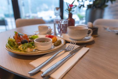 Salata, tütsülenmiş somon, zeytin, kahve ve suyla dolu bir yemek masası. Güzel çatal bıçak takımı ve kırmızı çiçekle süslenmiş, açık hava manzaralı..
