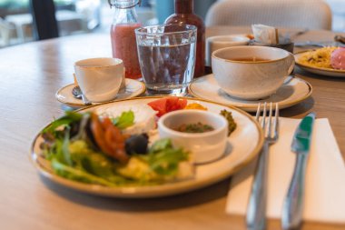Yeşiller, domatesler, zeytinler, kahve, meyve suyu ve suyla dolu bir kahvaltı masası. Parlak, modern bir otel yemek alanında geniş pencereleri ve doğal ışığı olan bir yer..