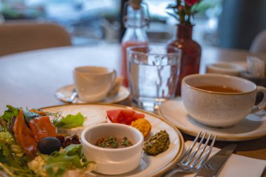 Lüks bir otelin yemek salonunda tütsülenmiş somon, yeşillik, zeytin, çay ve meyve suyu seti. Geniş pencereler açık hava manzarası sunar.