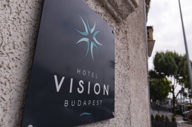 Duvarında deniz mavisi amblemi olan Budapeşte Oteli 'ne imza atın. Ağaçlara, sokağa ve bulutlu gökyüzünün altındaki binalara yaslanın..