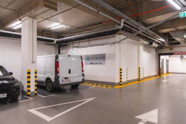 Yeraltı otoparkı, beyaz bir minibüs ve siyah bir araba, sarı güvenlik işaretleri, açık borular, parlak aydınlatma ve resepsiyon yönü için bir işaret..