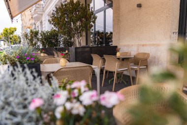 Açık hava oturma alanı bej hasır sandalyeler, masalar, saksı bitkileri ve ahşap bir güvertede çiçekler. Bej kaplama duvar ve cam pencereler boşluğu çerçeveler.