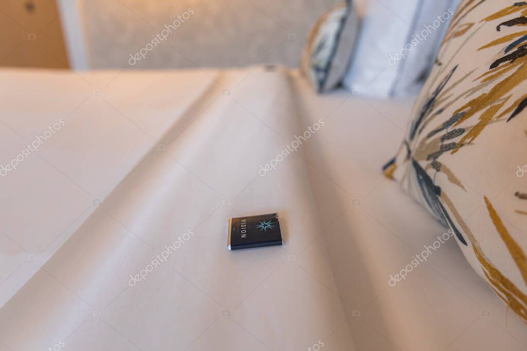 A luxury hotel bed with white linens, botanical patterned pillows, and a branded chocolate. A softly lit textured wall enhances the ambiance.