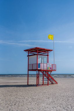 Ayaklıklar üzerinde kırmızı ve beyaz bir cankurtaran kulesi Parnu, Estonya 'da kumlu bir plajda duruyor. Arka planda sarı bir bayrak, durgun bir deniz ve açık mavi gökyüzü var..
