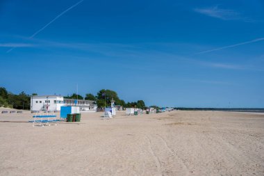 A serene beach in Parnu, Estonia, featuring a white building, lifeguard stands, and greenery under a clear blue sky. The coastline stretches into the distance.