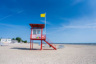 Estonya, Parnu 'da kumlu bir sahilde sarı bayrağı olan kırmızı bir cankurtaran kulesi duruyor. Salon sandalyeleri, şemsiyeler ve yakınlardaki küçük beyaz binalar görülebilir..