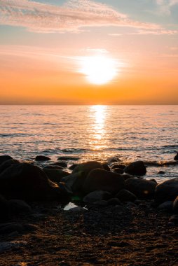 Estonya 'nın Parnu kentindeki Baltık Denizi üzerinde sakin bir günbatımı sıcak turuncu renkler, durgun su yansımaları, kayalık bir kıyı şeridi ve gökyüzünde ince bulutlar..