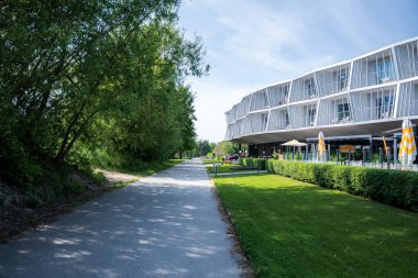 A uniquely designed white building with balconies, outdoor seating, and umbrellas, surrounded by greenery and a paved pathway under a clear blue sky.