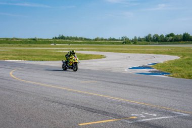 Sarı ve siyah dişli bir motosikletçi Porsche pistinde bir viraja giriyor, çimenli tarlalar, seyrek ağaçlar ve açık mavi bir gökyüzü ile çevrili..