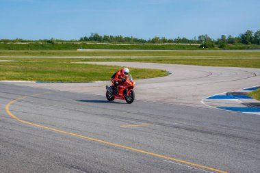 Kırmızı takım elbiseli ve kasklı bir motosikletçi kaldırımlı bir pistin kıvrımına eğiliyor. Üzerinde açık işaretler, çimenli bir çevre ve parlak mavi bir gökyüzü var..