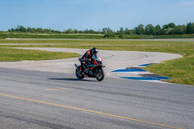 Tam yarış vitesli bir motosikletçi, asfalt, çimenlik alanlar, ağaçlar ve parlak mavi gökyüzü ile çevrili Porsche pistinde bir viraja doğru eğilir..