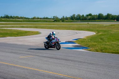 Tam teşekküllü yarış teçhizatlı bir motosikletçi mavi ve beyaz kaldırımlı bir pistte çim, ağaç ve açık mavi gökyüzü ile çevrili bir virajda yol alır..