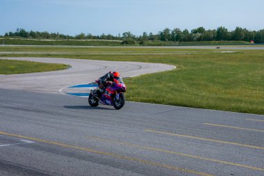Bir motosikletçi asfalt pistte kırmızı kask ve yarış kıyafeti giymiş bir viraja giriyor. Raylar çimenli tarlalar ve açık bir gökyüzünün altındaki ağaçlarla çevrili..