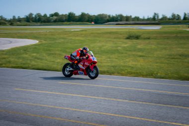 A red sports bike with number 224 races on a paved track surrounded by grass and trees. The rider wears a red and black suit, leaning forward.
