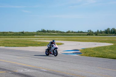 Tam yarış kıyafeti giymiş bir motosikletçi, Porsche pistinde çıkartmalı bir spor bisikleti sürüyor. Pürüzsüz asfalt, şerit işaretleri ve çimenli bir çevresi var..