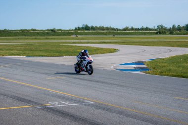 Bir motosikletli tam yarış kıyafeti ile dolambaçlı asfalt bir pistte otlar, seyrek ağaçlar ve açık mavi bir gökyüzü ile çevrili, mavi ve beyaz kaldırımlı.