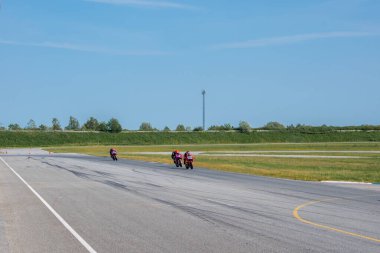 Açık bir gökyüzünün altında, çimenler ve ağaçlarla çevrili asfalt bir pistte üç spor bisikleti yarışıyor. Arka planda uzun bir iletişim kulesi duruyor..