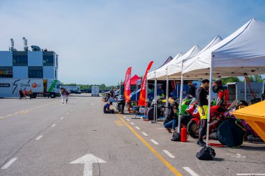 Porsche pistinde canlı bir bisiklet yarışı sahnesi, beyaz çadırların, renkli bisikletlerin, afişlerin, modern bir binanın ve açık asfaltın yer aldığı..