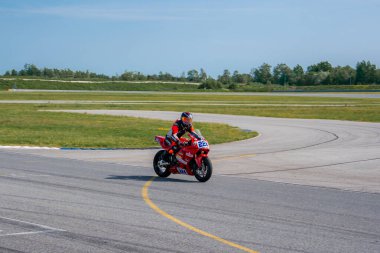 A motorcyclist in red and black gear rides a red bike marked 22 on a paved track with sharp curves, grassy surroundings, and a tree lined backdrop.