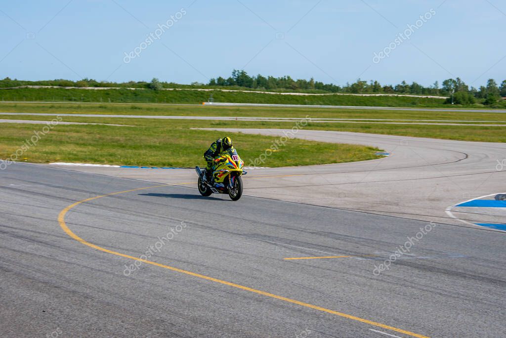 A motorcyclist in a yellow and black suit races on a sports bike at a track with sharp curves, grassy areas, and trees under a clear blue sky.