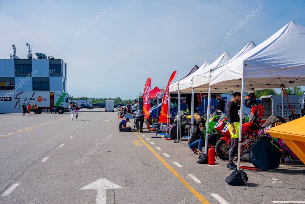 Vibrant scene of a sports bike racing event on the Porsche track, featuring white tents, colorful bikes, banners, a modern building, and open asphalt.