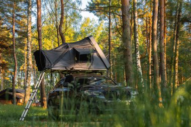 Katlanabilir çatı çadırı ve merdiveni olan modern bir SUV ormanda park halinde. Güneş ışığı, arka planda ahşap bir yapı bulunan uzun ağaçların arasından süzülür..