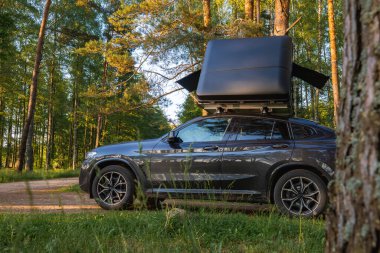 Çadırı kapalı modern bir SUV yemyeşil bir ormanda kirli bir yola park edilmiştir. Güneş ışığı, uzun yeşil ağaçların arasından süzülüp huzur verici bir sahne yaratır..