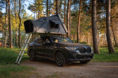 Çadırı katlanabilir bir çadırı olan modern bir SUV, ormandaki kirli bir yola park edilmiş. Aracın etrafını saran uzun çam ağaçları güneş ışığıyla filtreleniyor..