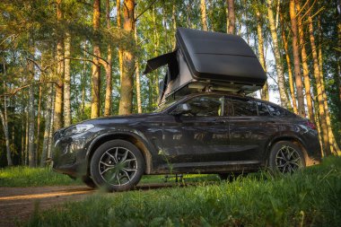 Kısmen açık çatı çadırına sahip gösterişli bir SUV yeşil yapraklı uzun ağaçların arasına park edilmiştir. Güneş ışığı filtreler ve sıcak bir atmosfer yaratır..