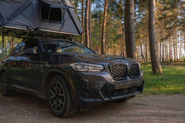 Yarı açık çatısı çadırı olan siyah bir SUV ormanda park halinde. Aracın etrafını saran uzun ağaçlar, gölgelikten süzülen güneş ışığı....