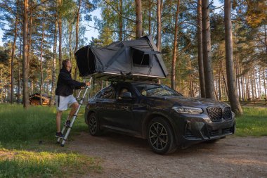 Bir adam, orman açıklıklarında park etmiş modern bir SUV 'un tepesindeki çadıra merdiven çıkıyor. Uzun çam ağaçları ve ahşap bir kulübe kırsal bir ortam yaratır..