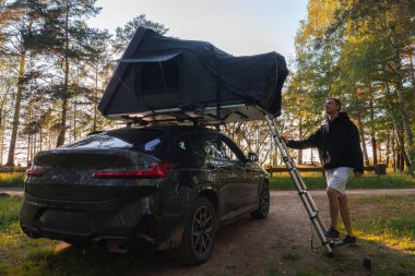 Ormana park edilmiş modern bir SUV, merdiveni olan bir çatı üstü çadırı. Uzun ağaçlar ve yumuşak güneş ışığıyla çevrili bir insan yakınlarda duruyor..