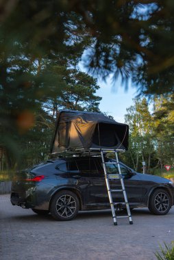 Uzun ağaçlar ve yeşilliklerle çevrili bir ormanda asfalt bir yüzeye park edilmiş katlanabilir çatı çadırı ve metal merdiveni olan modern bir SUV. Bir verim işareti görünür.