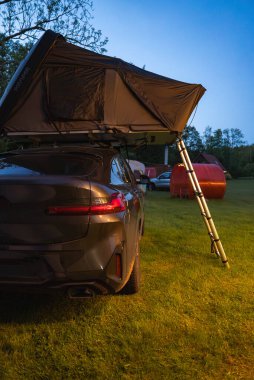 Günbatımında çimenli bir alana kurulmuş katlanabilir çatısı olan modern bir SUV. Arka planda yapılar ve ağaçlar gibi kırmızı kabin görünüyor..