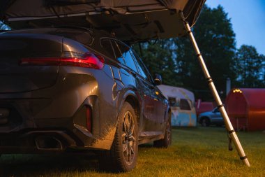 Ormandaki bir kamp alanının çimlerine park edilmiş katlanabilir çatı çadırına sahip modern bir SUV. Çamurlu detaylar, karavan ve ağaçlar dış mekanı güzelleştiriyor..