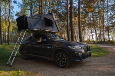 Katlanabilir çatı çadırı ve merdiveni olan siyah bir SUV ormanda park halinde. Dalların arasından süzülen uzun ağaçlar ve güneş ışığı huzurlu bir ortam yaratır..