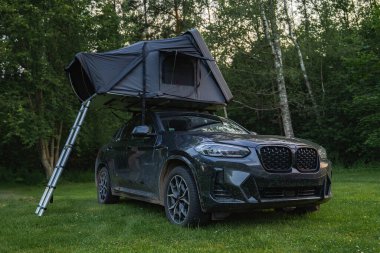 Çimenli bir ormanlık alana kurulmuş katlanabilir çatı çadırı olan modern bir SUV. Uzun yeşil ağaçlarla çevrili bir merdiven yere kadar uzanır..