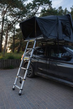 Ormanın asfalt bir yüzeyine park edilmiş modern bir SUV, teleskopik merdiveni olan siyah bir çatı üstü çadırı. Uzun ağaçlar ve güneş ışığı huzurlu bir ortam yaratır..