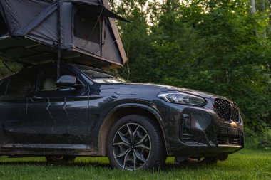 Kısmen açık çatı çadırına sahip modern bir SUV sık bir ormanda çimlerin üzerine park edilmiştir. Araç, doğa ile harmanlanmış, off-road yolculuğundan toprak gösteriyor..