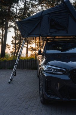 Çatı çadırı ve merdiveni olan modern bir SUV ormandaki ahşap bir bariyerin yanında park edilmiştir. Gün batımı ışık filtreleri arkaplandaki ağaçların arasından geçer.